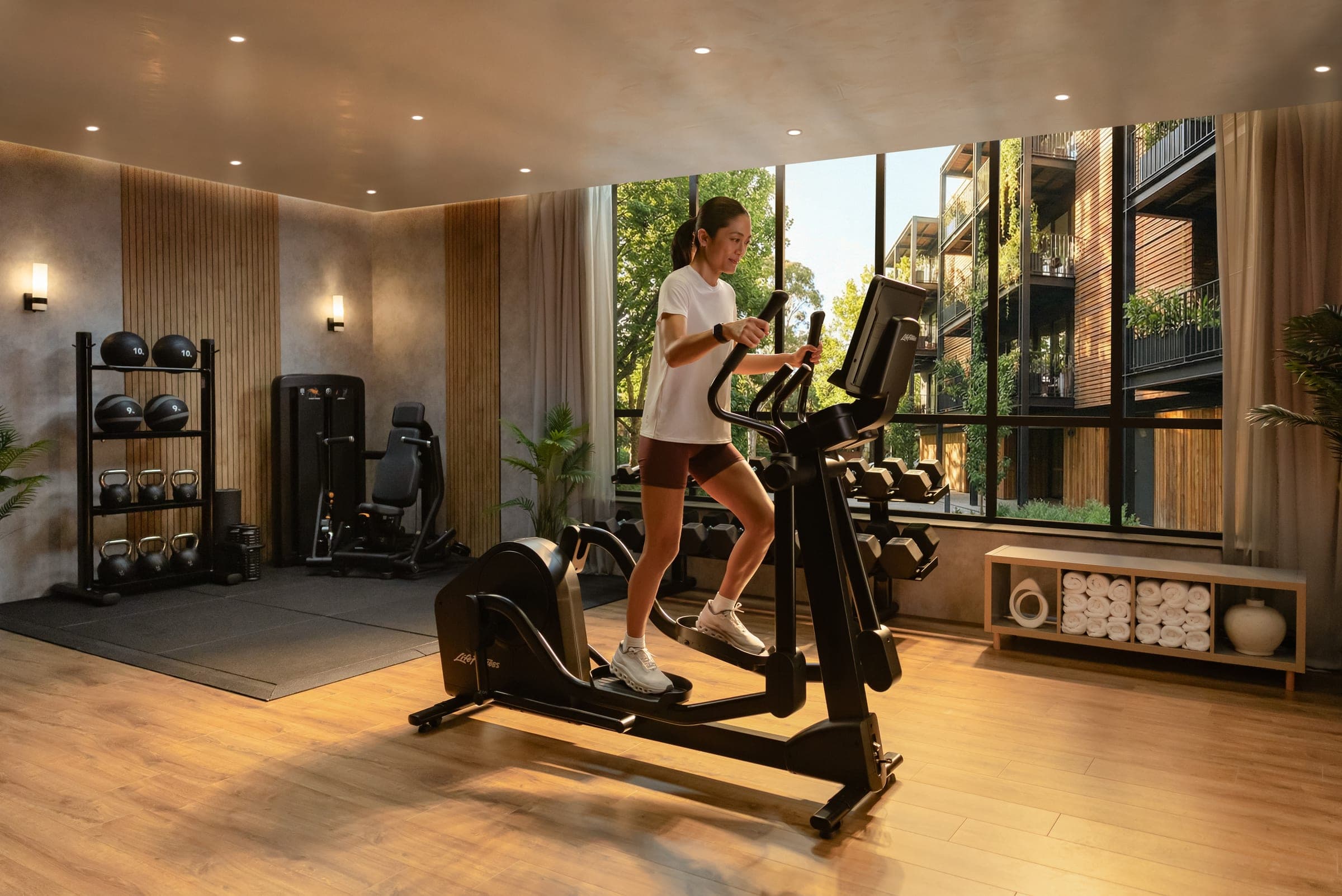 Fitness v bytovém domě s výhledem do zeleného dvora, eliptický trenažér a volné váhy